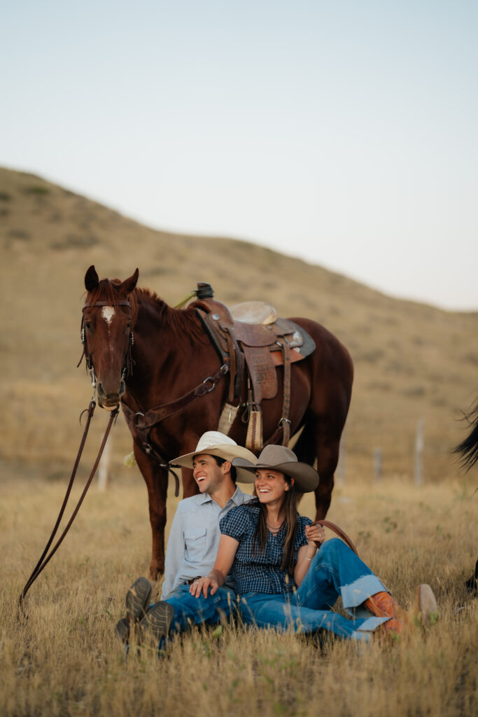 man and woman sitting and laughing with a horse behind them