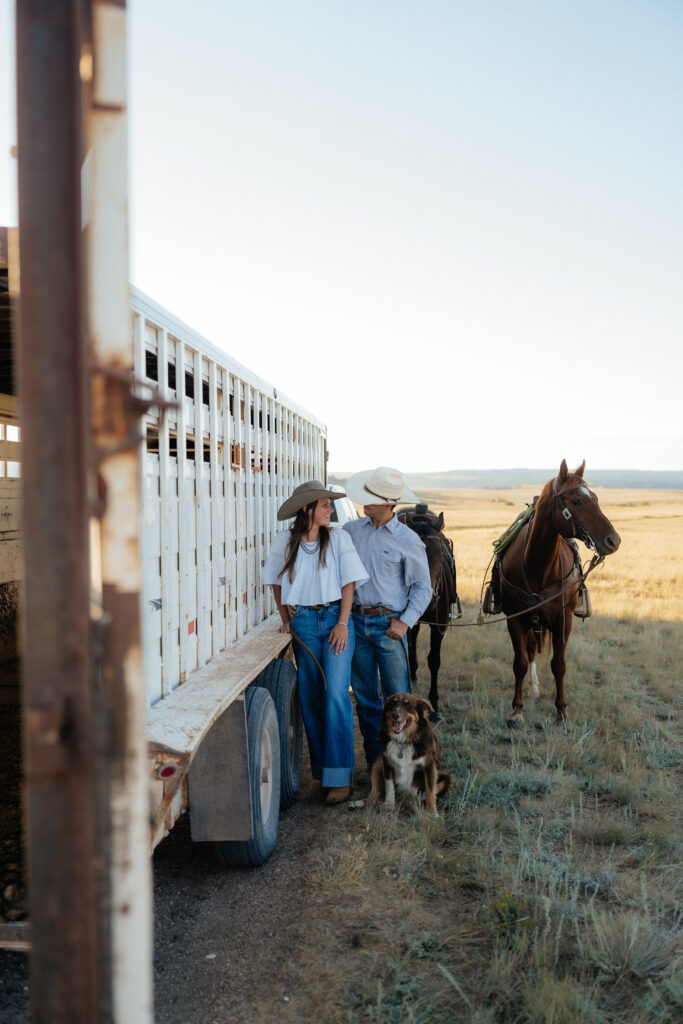 man and woman looking at one another while leaning against a trailer with their dog and horses